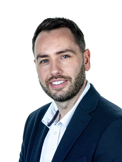A man with short dark hair and a beard, wearing a white shirt and dark blazer