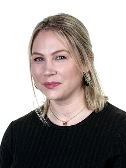 A woman with shoulder-length blonde hair, wearing a black top and gold necklaces, smiles softly