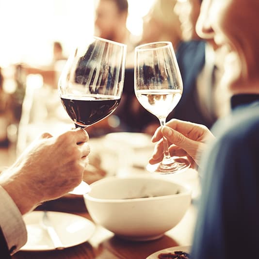 People clinking glasses of red and white wine in a warm, social setting with a bowl on the table.