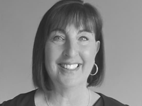 Smiling woman with short dark hair and hoop earrings, wearing a dark top, against a plain background. Black and white image.