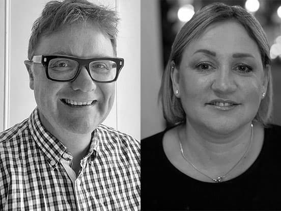 Black and white photo of a man in glasses wearing a checkered shirt and a woman with short hair wearing a dark top, both smiling.