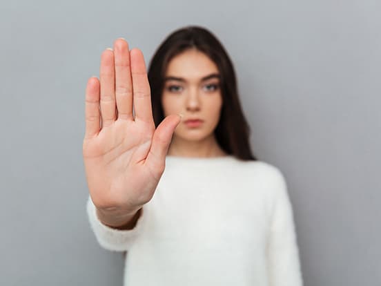 Woman in a white sweater holding her hand up toward the camera, palm open, against a gray background.