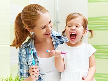 Mother smiling and helping young daughter brush her teeth
