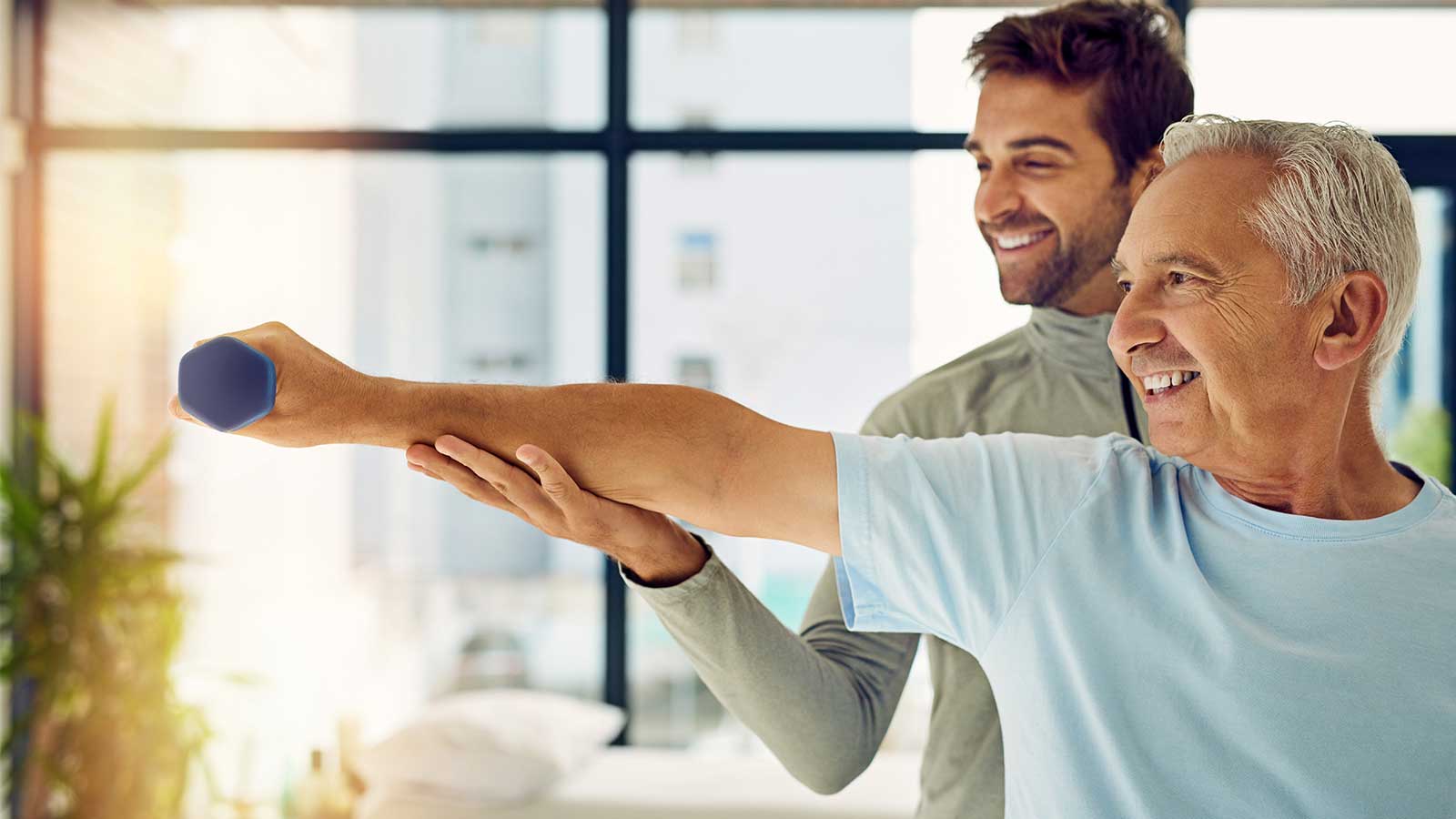 Elderly man exercises with a dumbbell, guided by a smiling trainer in a bright room with large windows.