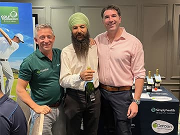 Three men smiling and posing together indoors, one holding a bottle. Golf-themed banners and a table with items are in the background.