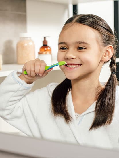 Young girl with pigtails wearing a white robe, smiling while brushing her teeth with a green toothbrush in a bathroom.