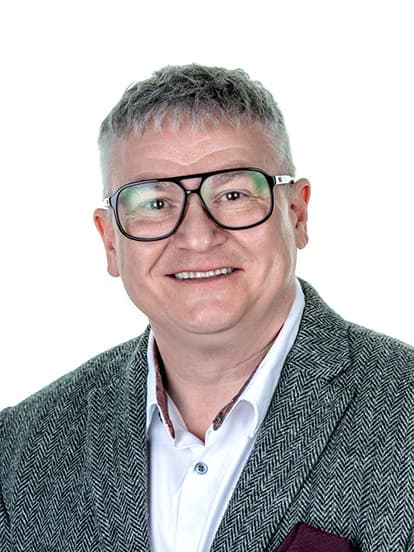 Smiling man with glasses and gray hair, wearing a herringbone blazer and white shirt