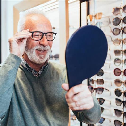 Elderly man trying on glasses, smiling while holding a hand mirror in an eyewear store with a display of sunglasses in the background.