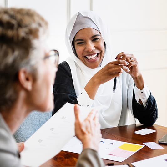 Two women sitting at a table, smiling and talking. One is holding papers, and the other is wearing a hijab and holding a pen.