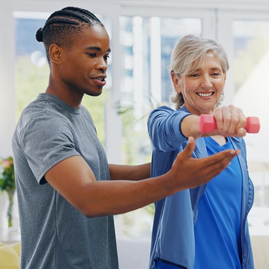 A woman exercises with a pink dumbbell, guided by a trainer in a grey shirt, in a bright room.