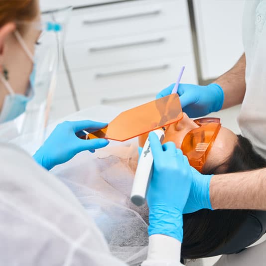 Dentist and assistant performing a dental procedure on a patient wearing protective glasses, using a UV light tool and orange shield.