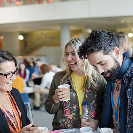 Three people are standing together, smiling and holding cups at a social event. They appear to be engaging in a pleasant conversation.