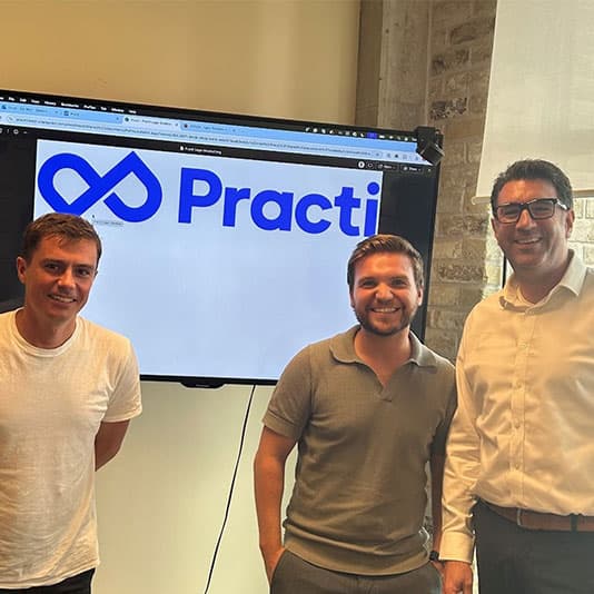 Three men stand smiling in front of a screen displaying the Practi logo in an office setting.