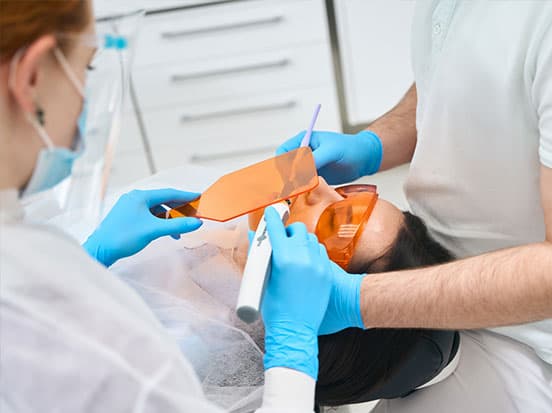 Dentist and assistant performing a dental procedure on a patient wearing protective eyewear, using dental tools in a clinical setting.