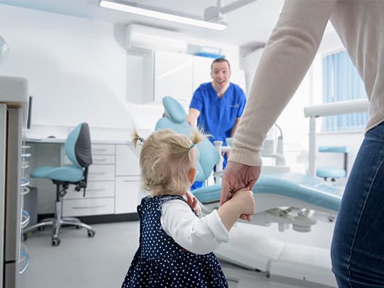 A toddler holding an adult's hand enters a dental clinic, greeted by a smiling dentist in blue scrubs.